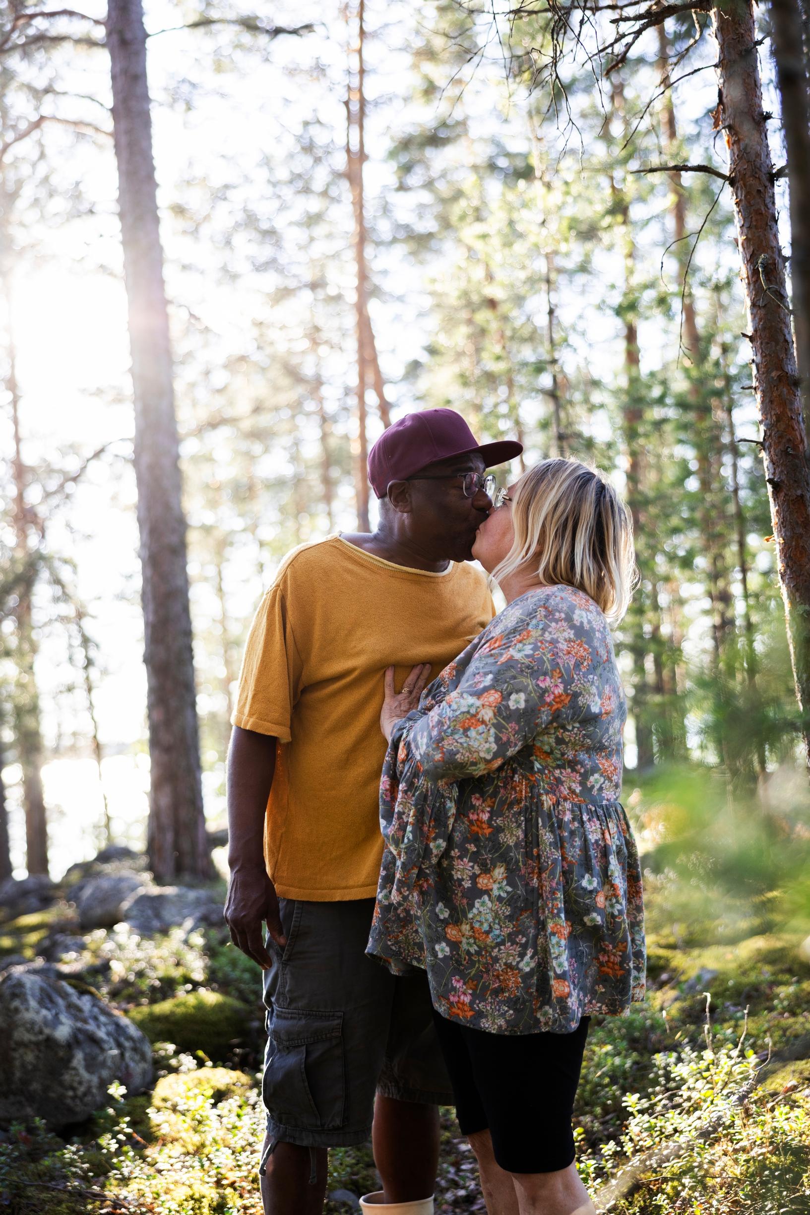 lippalakkipäinen mies ja kukkapaitainen nainen suutelevat kesäisessä metsässä