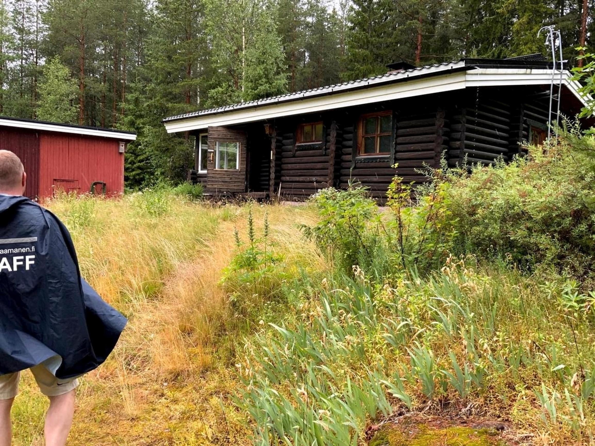 sadeviittaan pukeutunut mies kävelee heinikkoa kohti tummaa hirsimökkiä ja punaista rakennusta