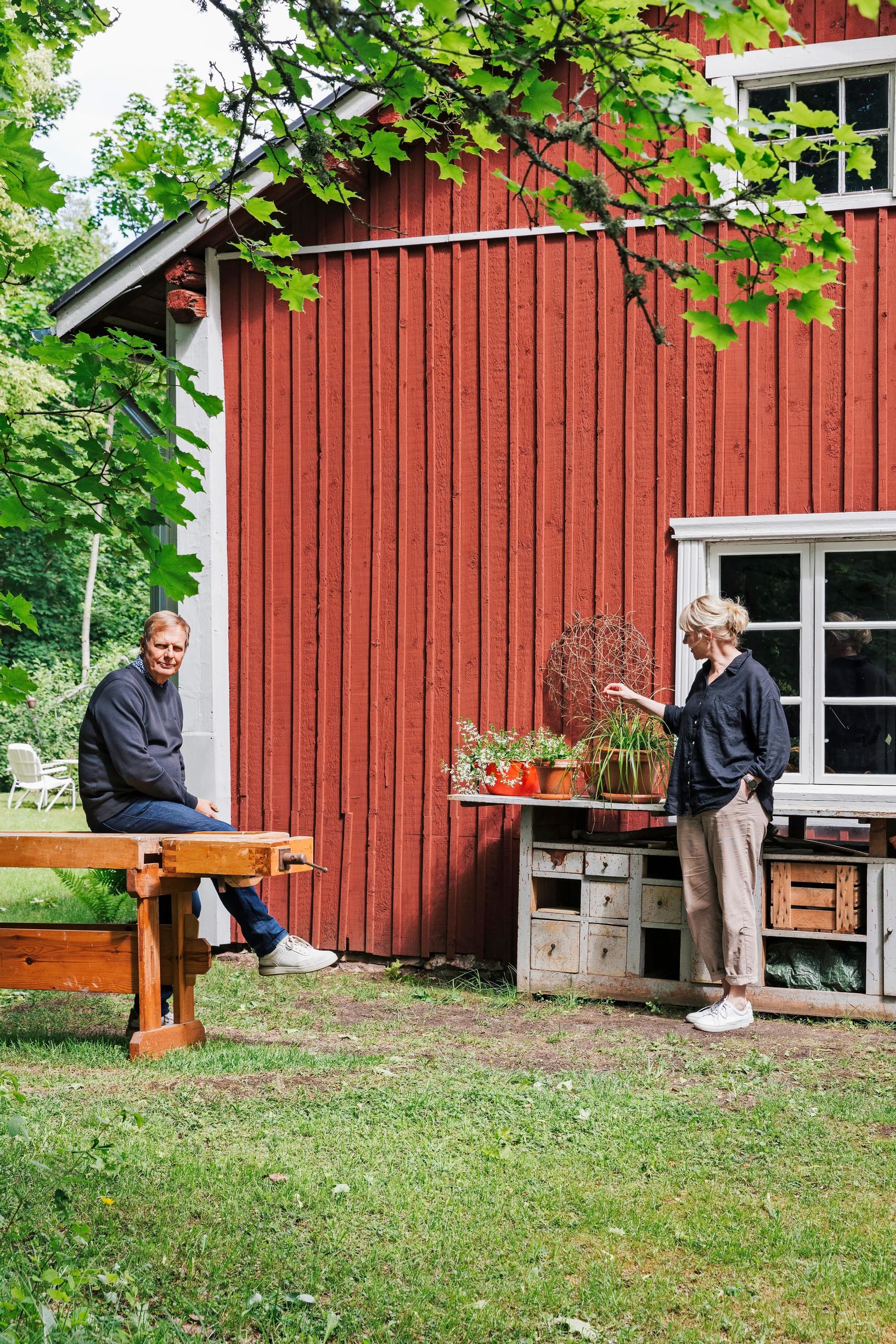 Anne Pakomäki ja Heikki Hyttinen pihalla, Heikki istuu höyläpenkillä ja Anne seisoo talon vieressä