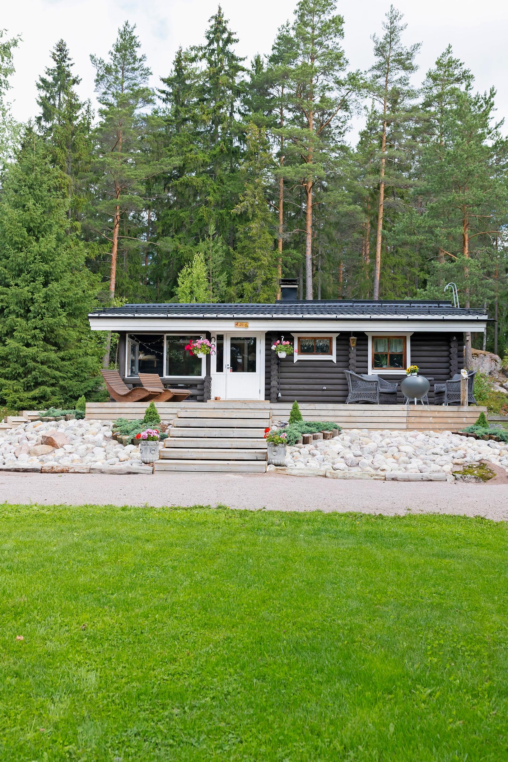 tumma mökki ja siisti miljöö, terassi ja kivikkorinne, portaat ja nurmikenttää