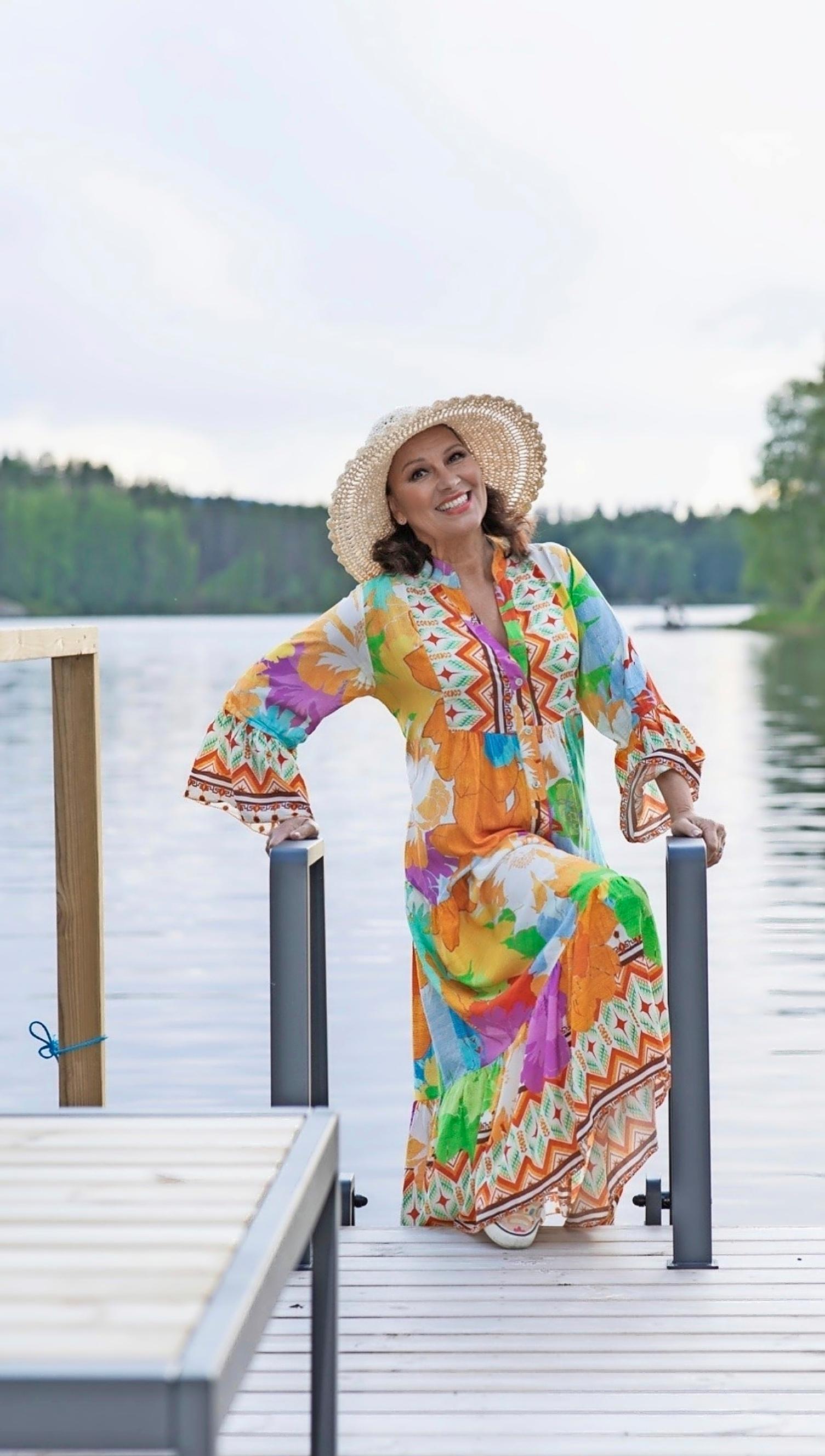 Arja Koriseva värikkäässä mekossa lierihattu päässä mökkilaiturilla