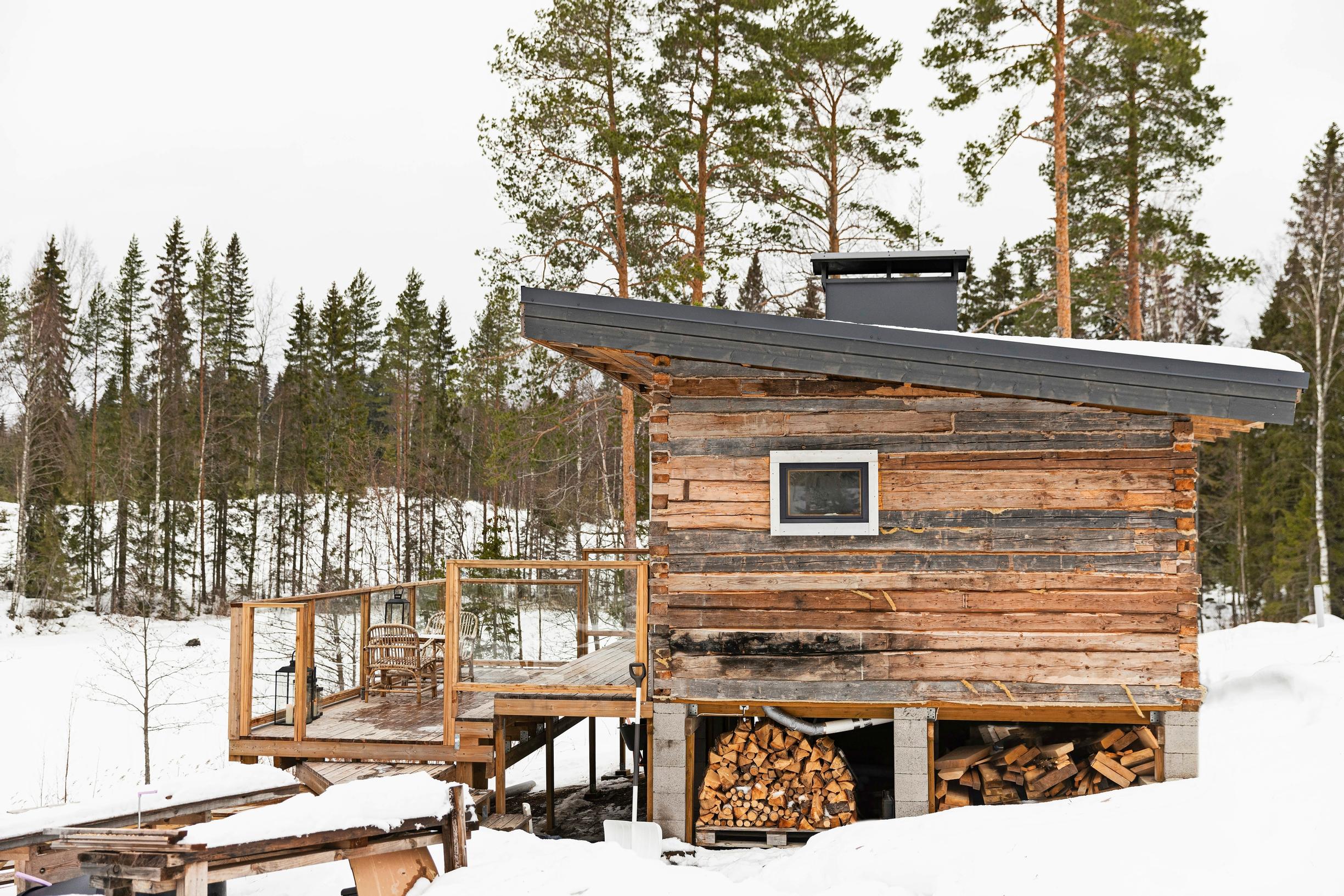 huonokuntoinen hirsirakennus lumisen järvenrannassa, taustalla metsää