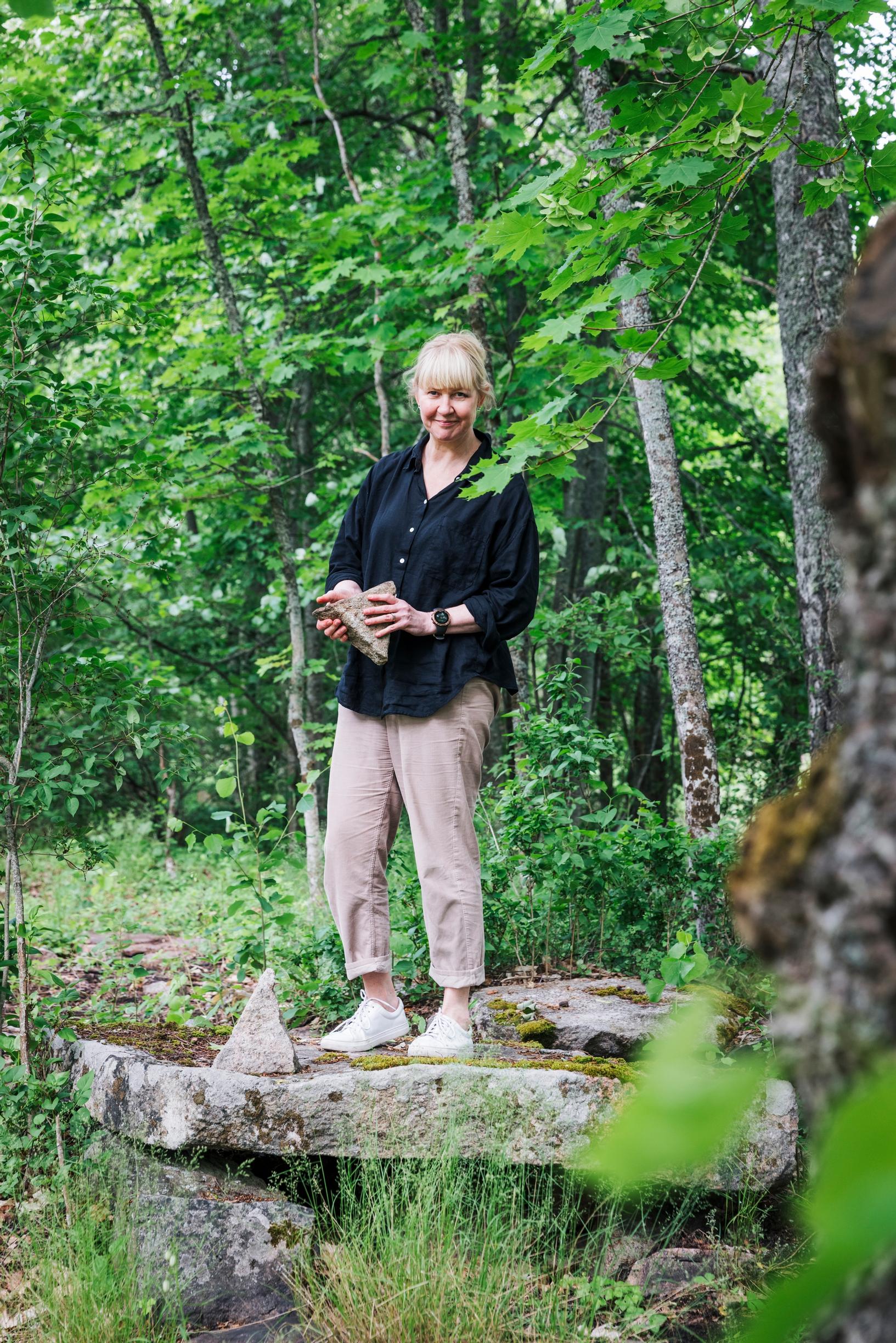 Anne Pakomäki seisoo vanhalla kivellä, rakennuksen perustuksella metsän reunassa