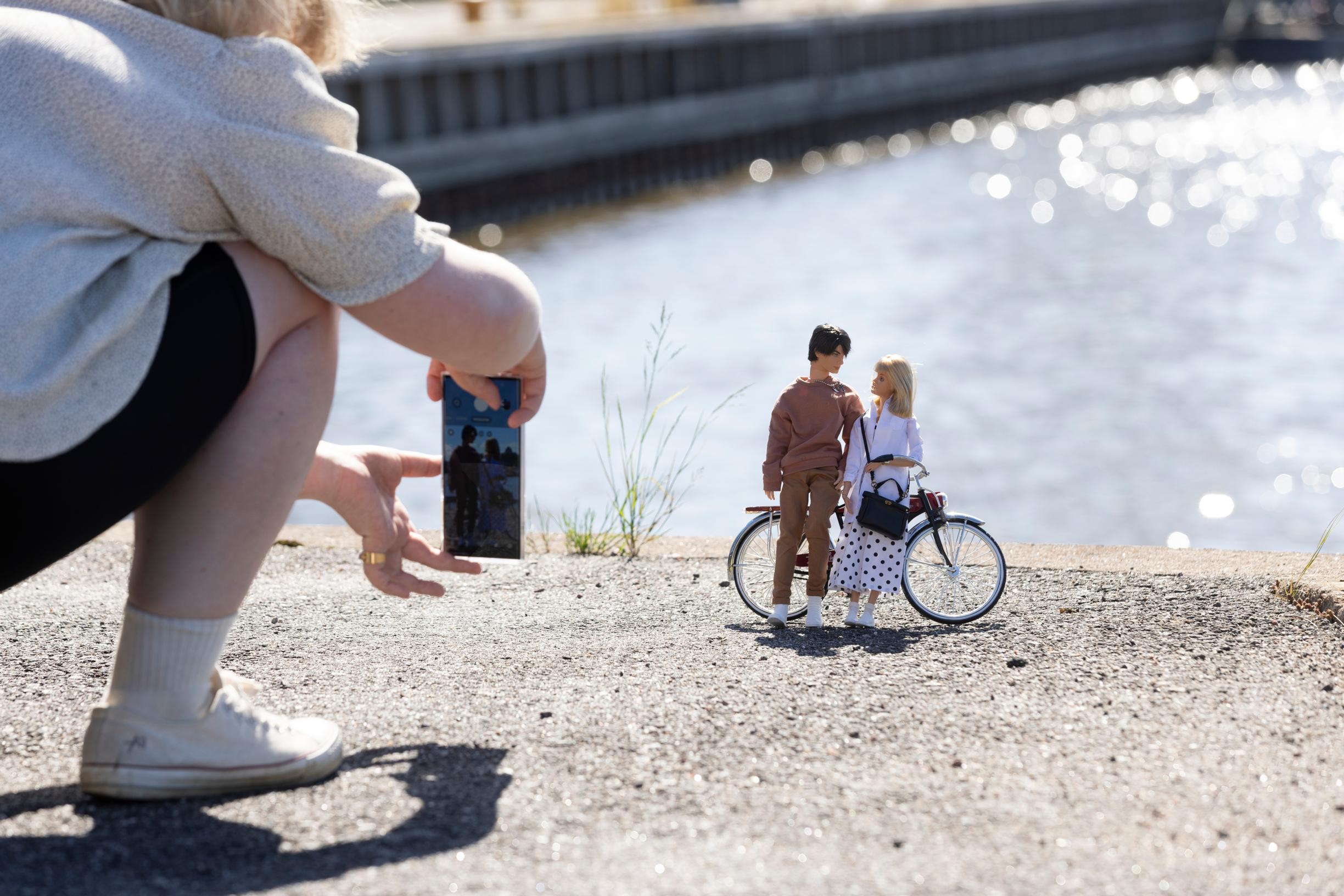 nainen ottaa kuvaa Barbie- ja Ken-nukeista, jotka nojaavat polkupyörään joen rannalla
