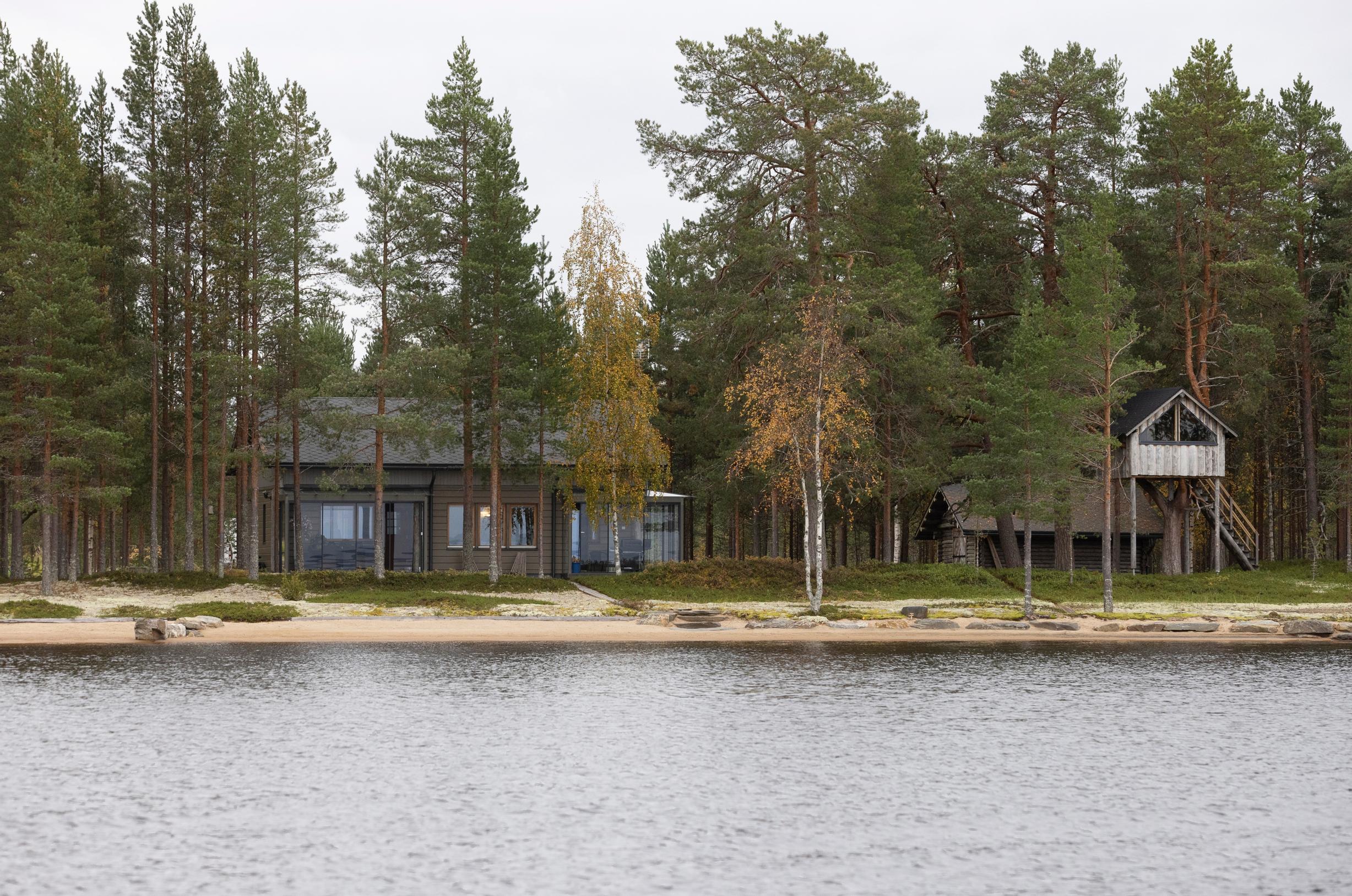 tumma mökki, toinen rakennus ja maja pylväillä järveltä päin katsottuna, metsää
