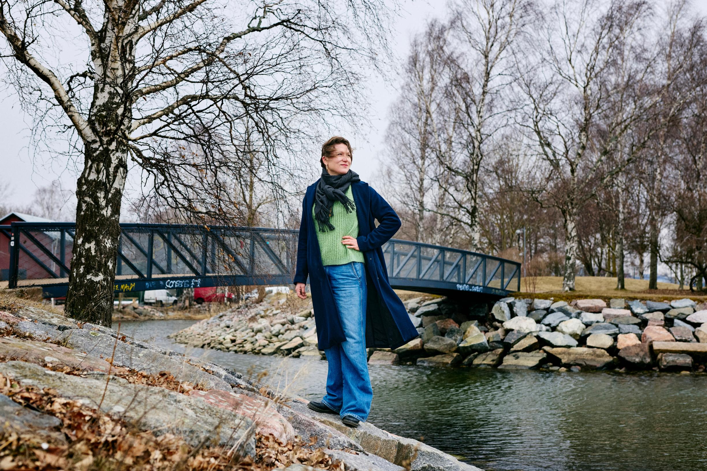 nainen seisoo joen varressa, taustalla näkyy silta ja koivuja