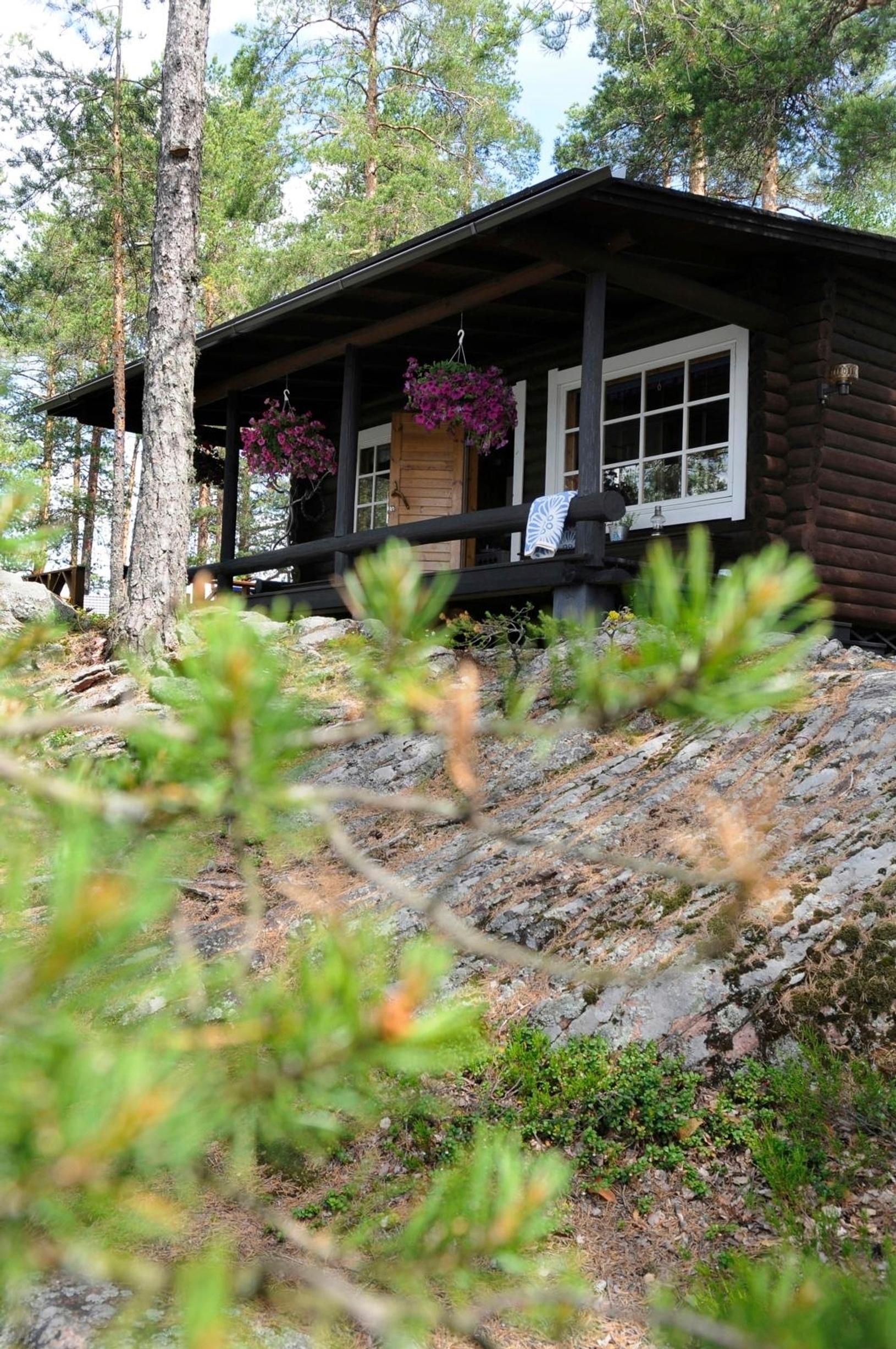musta hirsimökki mäellä, edessä kalliota ja ympärillä metsää
