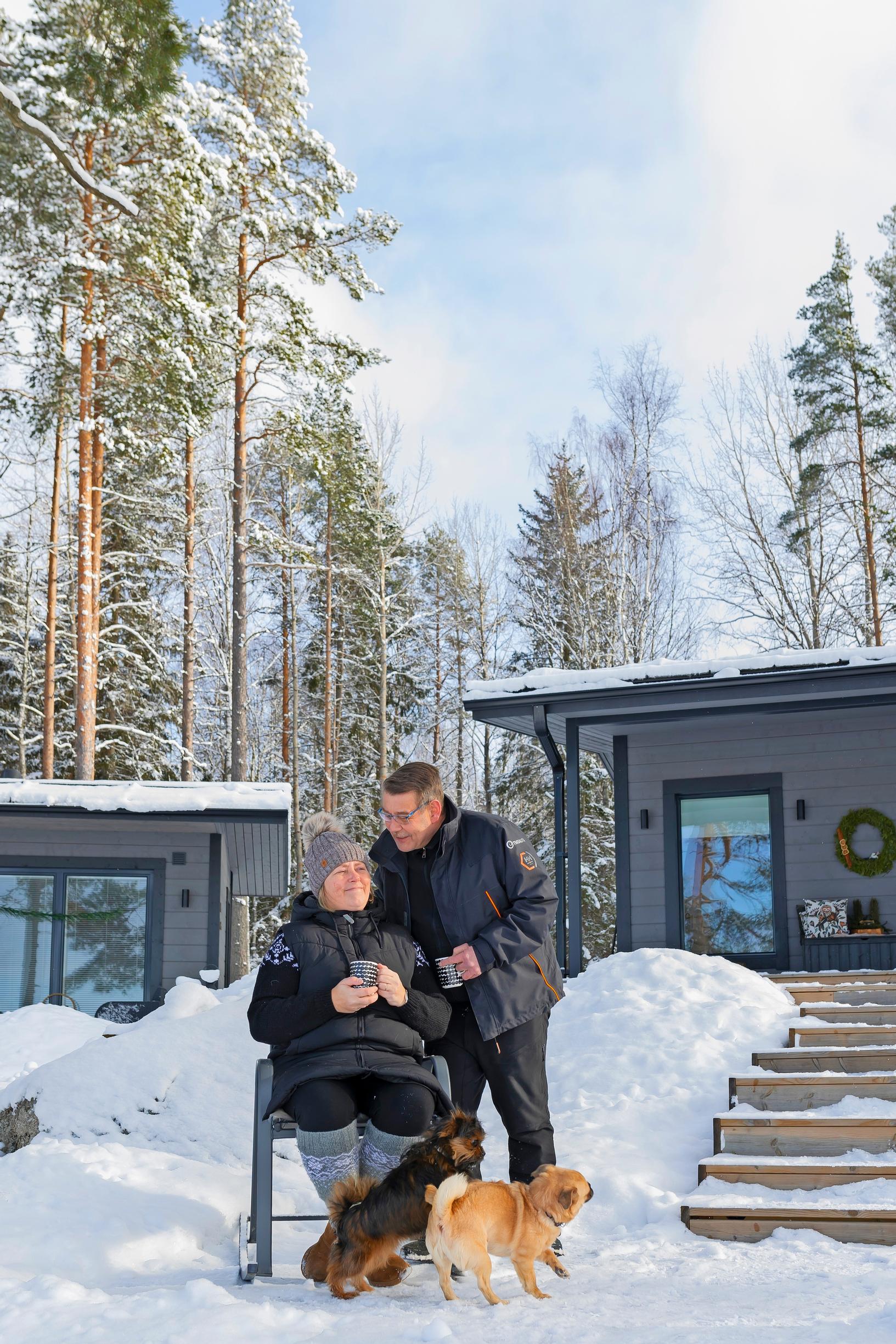 Mari istuu ja Kari mökkipihalla, kaksi koiraa ja taustalla kaksi rakennusta, lunta ja metsää