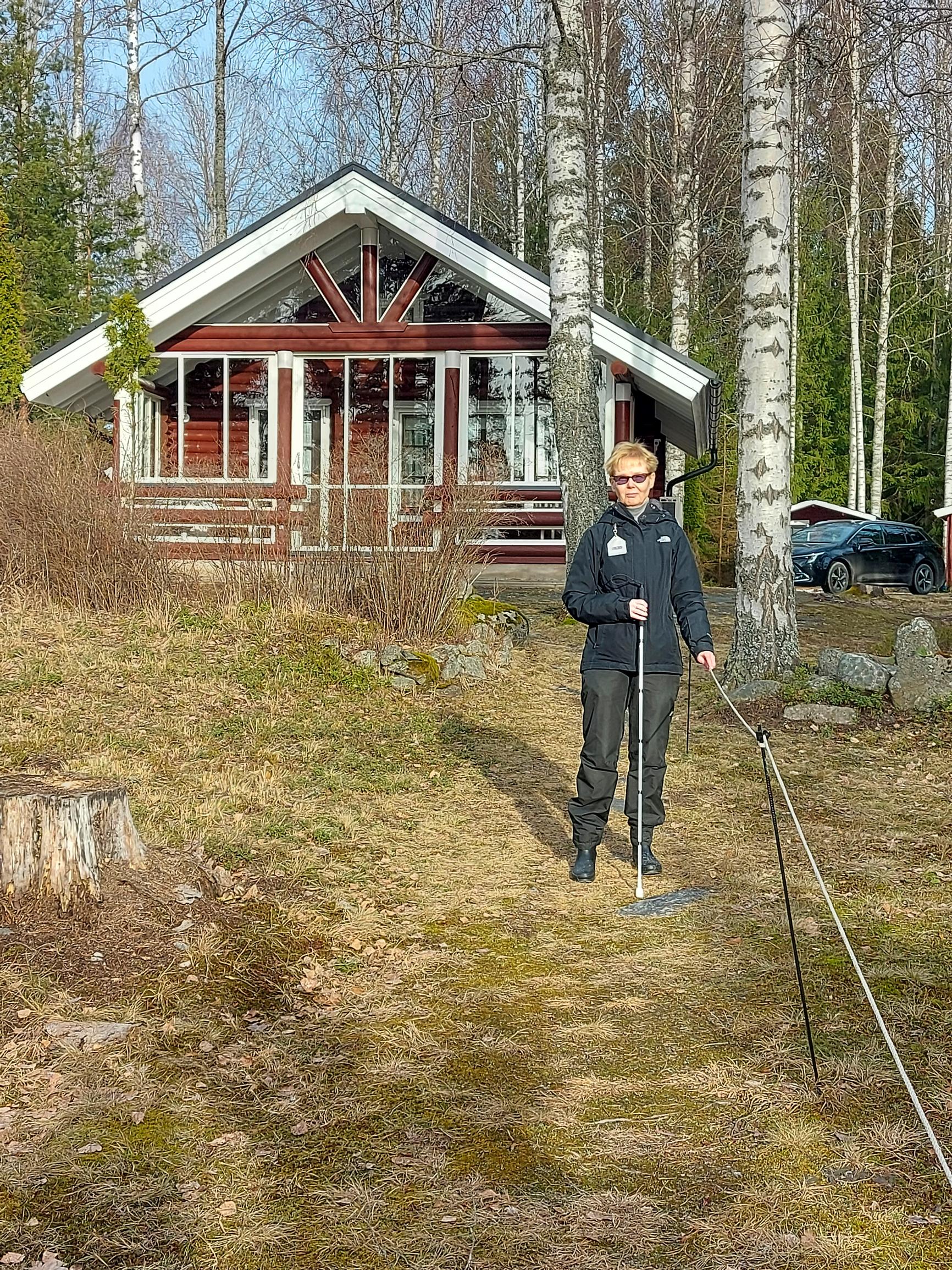 Merja Heikkonen pitää kiinni ohjausnarusta keppi toisessa kädessä, taustalla punainen kesämökki