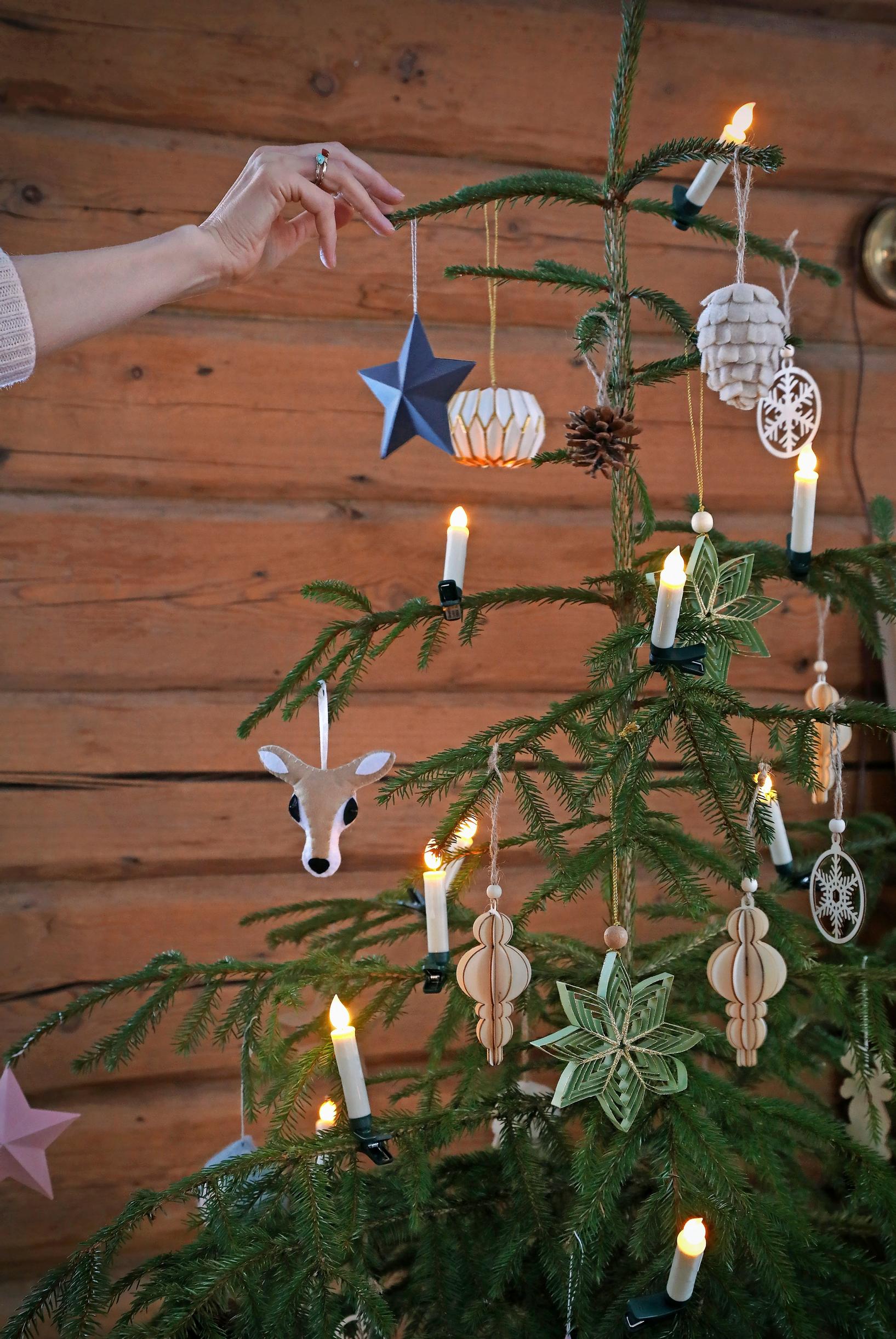 Ullan käsi ripustamassa joulukuuseen koristetta, taustalla hirsiseinä