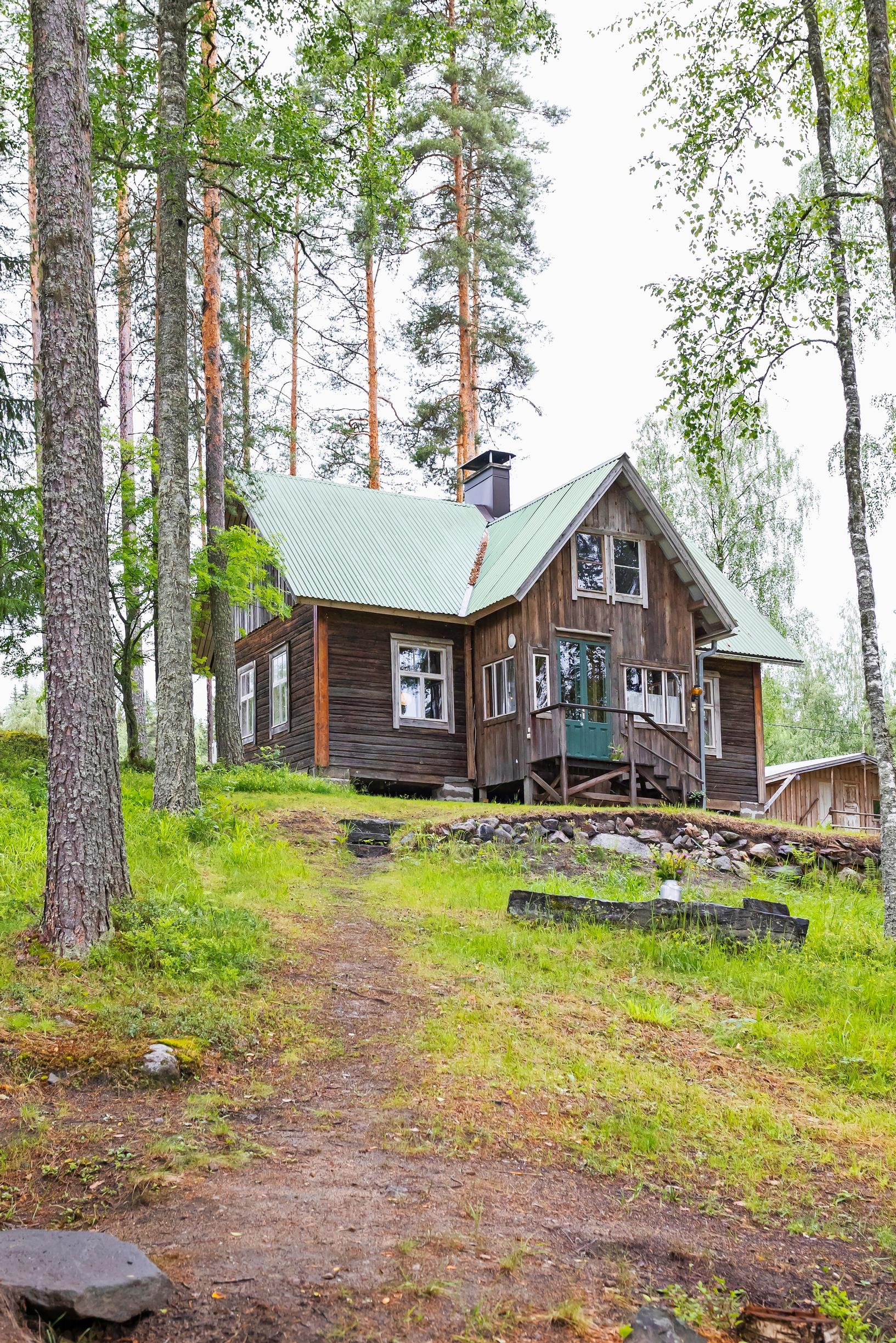 mäellä metsän reunassa harmaa vanha hirsimökki, jonka edessä kivimuuria ja ruohoa rinteessä